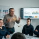 Leader che parla animatamente al team in riunione davanti a uno schermo con grafici in calo, tre collaboratori con filamenti dorati superficiali sulle spalle: una sorride compiaciuta, un giovane resta in silenzio stanco, un altro annuisce a braccia conserte
