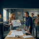 Ufficio tecnico in officina, presentazione di un organigramma al titolare mentre alcuni collaboratori seguono e altri restano chiusi nella propria postazione allineamento degli obiettivi